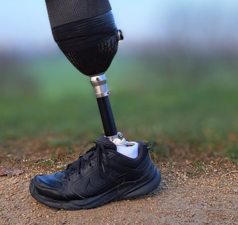 Close-up of a prosthetic leg wearing a black athletic shoe on a dirt path.